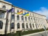 Gebäude des Bundesministeriums für Digitales und Verkehr bei Sonnenschein - vor dem Ministerium wehen die EU-, die deutsche und die ukrainische Flagge (v. l. n. r.). Auf dem Gehweg ist eine Person unterwegs.